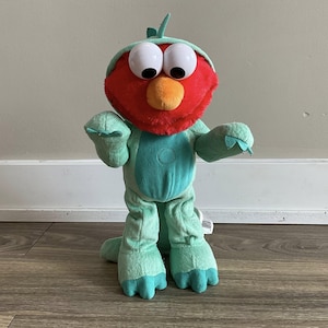 May include: A soft toy of Elmo from Sesame Street, costumed as the Statue of Liberty. Elmo has a red face, large white eyes, and an orange nose. He wears a light green outfit with a matching crown and has his arms outstretched.