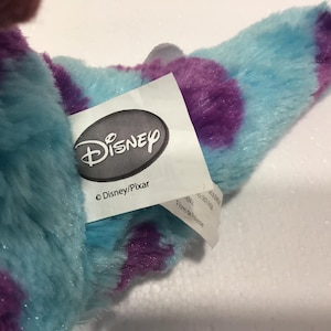 Op de afbeelding: Een blauw en paars pluche speelgoed met een Disney/Pixar-label eraan bevestigd. Het label heeft het Disney-logo en de tekst "Disney/Pixar".