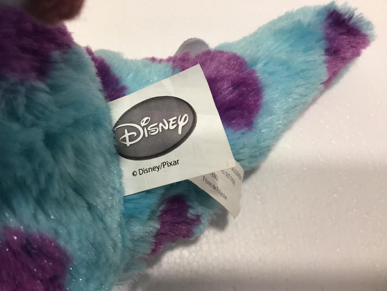Op de afbeelding: Een blauw en paars pluche speelgoed met een Disney/Pixar-label eraan bevestigd. Het label heeft het Disney-logo en de tekst "Disney/Pixar".