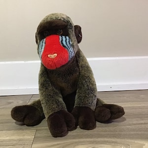 May include: A plush toy of a mandrill monkey with brown fur, a red face, and blue and white stripes on its nose. The monkey is sitting on a wooden floor.