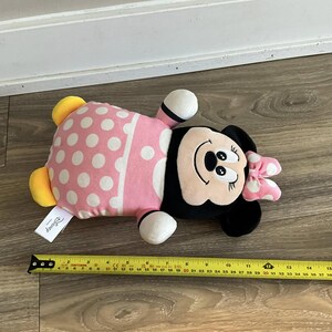 K&ouml;nnte beinhalten: Rosa-wei&szlig;es Minnie Mouse Pl&uuml;schtier mit schwarzen Punkten und einer schwarzen Schleife. Das Spielzeug liegt auf einem Holzboden mit einem Ma&szlig;band daneben. Das Spielzeug ist etwa 30 cm lang.