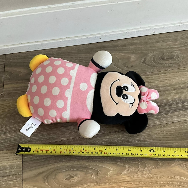 K&ouml;nnte beinhalten: Rosa-wei&szlig;es Minnie Mouse Pl&uuml;schtier mit schwarzen Punkten und einer schwarzen Schleife. Das Spielzeug liegt auf einem Holzboden mit einem Ma&szlig;band daneben. Das Spielzeug ist etwa 30 cm lang.