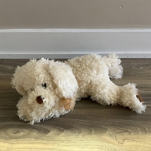 May include: A plush toy dog in a lying position. The dog is cream-colored with a shaggy texture, brown accents on the ears and nose, and black eyes. It is lying on a wooden floor.