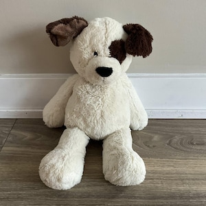 May include: A soft toy dog with a cream-coloured body, brown ears, and a brown spot over one eye. The dog has a black nose and is sitting upright. The toy is made of soft, fuzzy material.