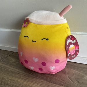 Könnte beinhalten: Ein gelbes und rosa Plüschtier in Form eines Boba-Tees mit einem weißen Deckel und einem rosa Strohhalm. Das Spielzeug hat ein lächelndes Gesicht mit geschlossenen Augen und rosa Herzen und Punkten auf der unteren Hälfte. Das Spielzeug hat ein Etikett mit der Aufschrift "Original Squishmallows" und "Bergit".