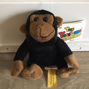 May include: A black plush monkey toy with brown fur on its face and hands. The monkey is sitting with its legs crossed. The toy has a tag that says "Tiny TossieMs Bean Filled Toy".