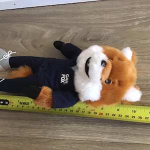 Pu&ograve; includere: Un peluche a forma di volpe con pelliccia arancione e bianca, che indossa una giacca blu navy con la scritta "CAR FOX" e pantaloni abbinati. La volpe &egrave; sdraiata, con scarpe da ginnastica grigie e una coda soffice. Il giocattolo &egrave; lungo circa 30 cm.