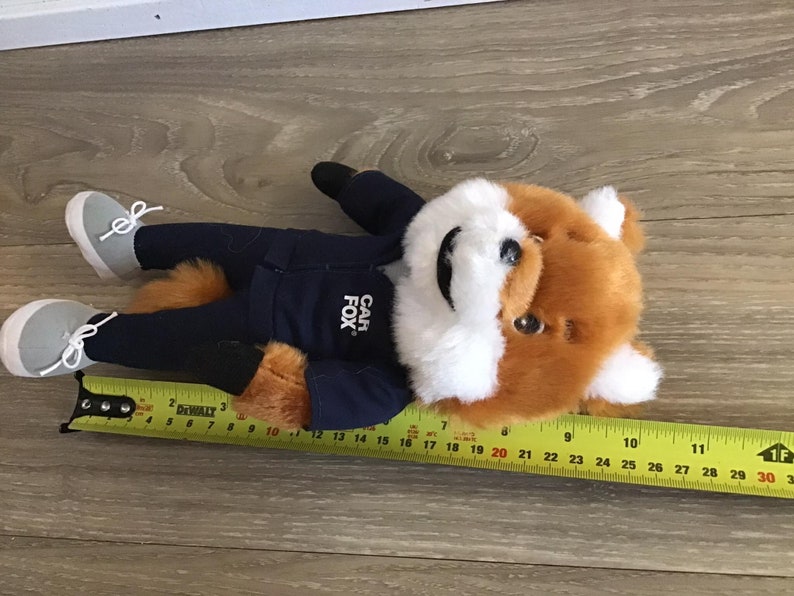 Pu&ograve; includere: Un peluche a forma di volpe con pelliccia arancione e bianca, che indossa una giacca blu navy con la scritta "CAR FOX" e pantaloni abbinati. La volpe &egrave; sdraiata, con scarpe da ginnastica grigie e una coda soffice. Il giocattolo &egrave; lungo circa 30 cm.