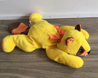 Könnte beinhalten: Ein gelbes Plüschtier mit braunen und orangen Akzenten. Das Spielzeug hat einen langen Schwanz und eine Schleife um den Hals gebunden. Das Spielzeug liegt auf einem Holzboden.
