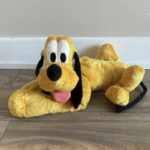 以下が含まれることがあります： 黒い耳と黒い尻尾を持つ黄色のぬいぐるみ犬。犬はピンクの舌を出していて、白い目があります。犬は横向きに寝ていて、頭は手の上に置かれています。
