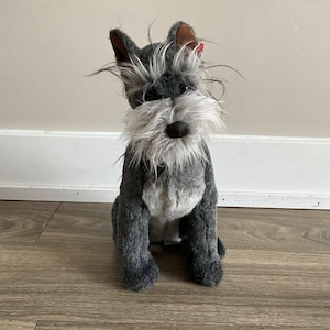 Op de afbeelding: Een pluche Schnauzer hondenspeelgoed in een zittende positie. Het speelgoed heeft een grijs en witte vacht, met lang, ruig haar rond het gezicht en een zwarte neus. De oren zijn bruin en het speelgoed zit op een houten vloer.