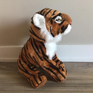 May include: A plush tiger toy with orange and black stripes, white fur on the chest and face, and black eyes. The toy is sitting on a wooden floor.