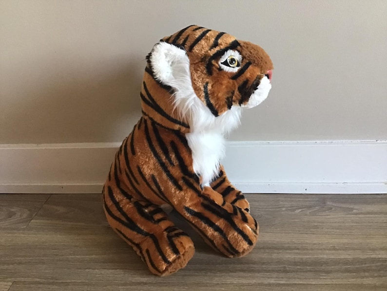 May include: A plush tiger toy with orange and black stripes, white fur on the chest and face, and black eyes. The toy is sitting on a wooden floor.
