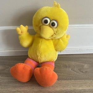 Puede incluir: Peluche amarillo Big Bird con pantalones a rayas naranja y rosa. El juguete tiene ojos grandes y redondos y un pico amarillo.