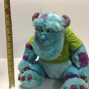 Op de afbeelding: Een blauw en paars pluche speelgoed van het personage Sulley uit de film Monsters Inc. Het speelgoed draagt een groen shirt en heeft een paarse patch op zijn arm. Het speelgoed is ongeveer 30 cm hoog.