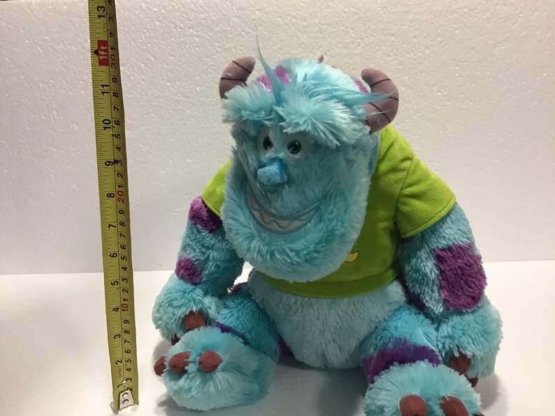 Op de afbeelding: Een blauw en paars pluche speelgoed van het personage Sulley uit de film Monsters Inc. Het speelgoed draagt een groen shirt en heeft een paarse patch op zijn arm. Het speelgoed is ongeveer 30 cm hoog.