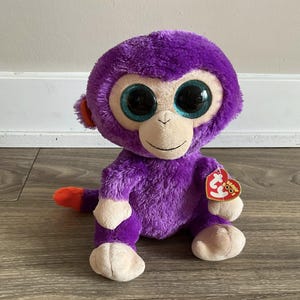 May include: A purple plush monkey toy with large, round, blue-green eyes and a tan face. The monkey has a red tail tip and ears, and a red heart-shaped tag. The toy is sitting on a wooden surface.