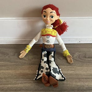 May include: A Jessie doll from the Toy Story franchise. The doll has red yarn hair, a white shirt with yellow and red accents, and cow-print trousers. The doll's brown boots have "ANDY" written on the soles.