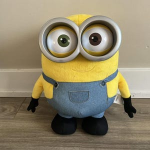 May include: A plush toy character with bright yellow skin, wearing blue denim-textured overalls and black gloves and shoes. The character features two large, prominent white eyes with silver-colored circular frames, one green iris and one brown iris.
