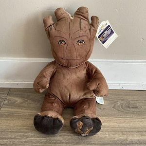 May include: A brown plush toy of the character Groot from the Guardians of the Galaxy. The toy has a tree-like appearance with a smiling face, large feet, and a tag from Build-A-Bear Workshop.