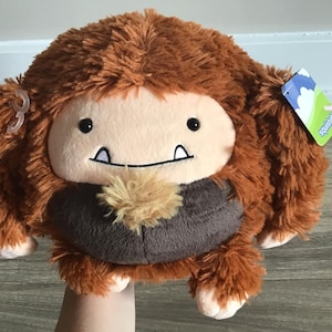 Puede incluir: Un juguete de peluche marrón y beige con textura peluda. El juguete tiene una cara sonriente con dientes blancos y un pequeño mechón de pelo marrón en el pecho. El juguete tiene una etiqueta que dice "Squishable".