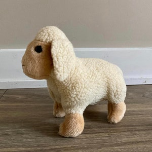 Może przedstawiać: Pluszowa zabawka w kształcie białej owcy z brązową twarzą i czarnymi oczami. Owca stoi na drewnianej podłodze.