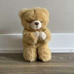 Pode incluir: Um urso de peluche marrom com os olhos fechados está rezando com as patas juntas. O urso está sentado em um chão de madeira.