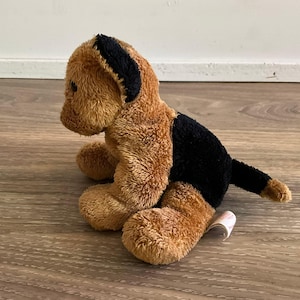 May include: A plush toy shaped like a dog with tan fur on the head, chest, and legs, and black fur on the ears, back, and tail. The stuffed animal is in a seated position with a small white fabric tag visible near the rear leg area.