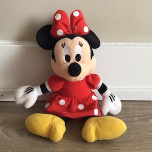 May include: A red and white polka dot dress with a red bow on top of the head. The plush toy has black eyes and a black nose. The toy is sitting on a wooden floor.