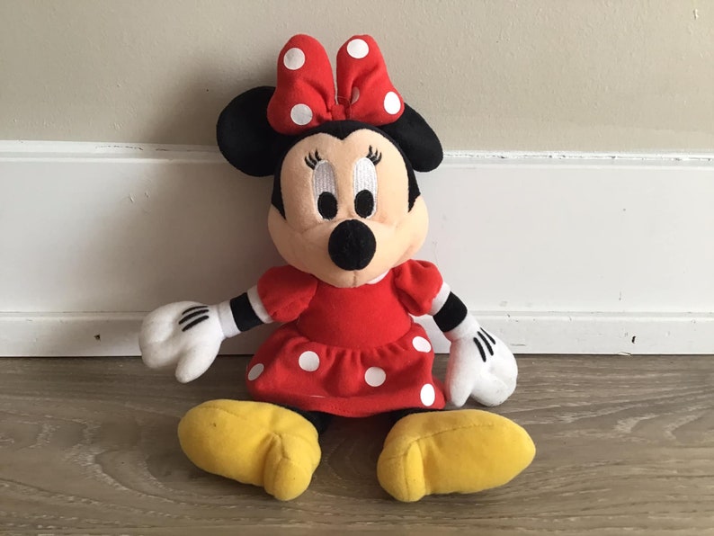 May include: A red and white polka dot dress with a red bow on top of the head. The plush toy has black eyes and a black nose. The toy is sitting on a wooden floor.