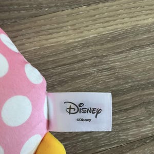 K&ouml;nnte beinhalten: Rosa-wei&szlig; gepunkteter Stoff mit einem gelben Stoffdetail. Der Stoff hat ein wei&szlig;es Etikett mit der Aufschrift "Disney" mit einem Copyright-Symbol und "Disney" darunter.