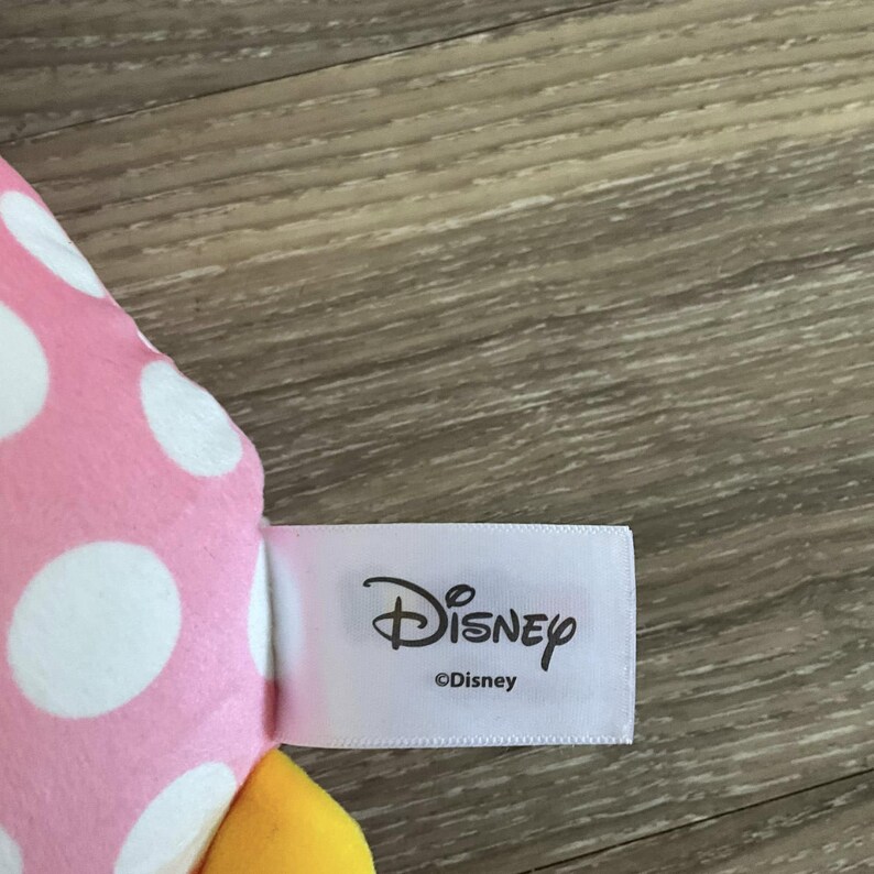 K&ouml;nnte beinhalten: Rosa-wei&szlig; gepunkteter Stoff mit einem gelben Stoffdetail. Der Stoff hat ein wei&szlig;es Etikett mit der Aufschrift "Disney" mit einem Copyright-Symbol und "Disney" darunter.