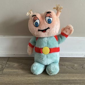 Può includere: Un giocattolo di peluche vintage a forma di alieno con testa rosa, occhi blu e bocca sorridente. Indossa una tuta azzurra con dettagli rossi e una fibbia gialla. Il giocattolo ha piccoli dettagli a forma di fiore sulla testa.