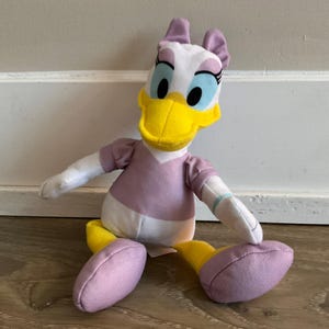 Könnte beinhalten: Ein Plüschtier in Form einer Ente mit weißem Gefieder, einem leuchtend gelben Schnabel und violetten Akzenten. Die Figur trägt ein violettes Oberteil mit Puffärmeln, violette Schuhe und hat violette Schleifen auf dem Kopf. Die Augen sind groß.