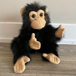 May include: A plush chimpanzee toy with black fur and a tan face, hands, and feet. The soft toy has large, round eyes and is posed with its hands up. The toy is designed to sit upright.