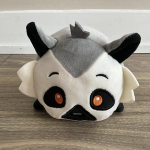 May include: A plush toy lemur with a white face, gray and black accents, and orange eyes. The lemur has large ears and a small, pointed crown. The toy is lying on a light-colored surface.