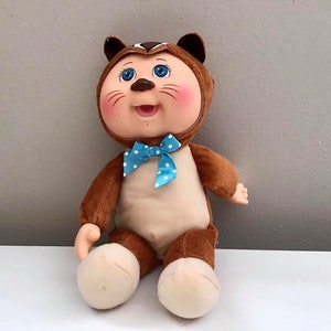 May include: A plush toy with a brown and white squirrel costume. The toy has blue eyes, a pink nose, and a blue bow tie with white polka dots.