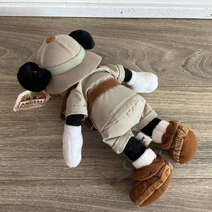 May include: A plush Mickey Mouse toy dressed in a tan safari outfit, including a matching pith helmet with a light green band, a brown belt, and brown boots. The toy features white gloves and a tag labeled Disney Animal Kingdom attached to the hat.