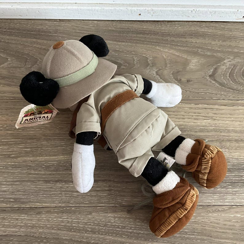 May include: A plush Mickey Mouse toy dressed in a tan safari outfit, including a matching pith helmet with a light green band, a brown belt, and brown boots. The toy features white gloves and a tag labeled Disney Animal Kingdom attached to the hat.