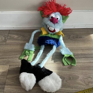 May include: A blue and green puppet with red hair, a yellow collar, and black shoes. The puppet has a large, toothy grin and is lying on a wooden floor.