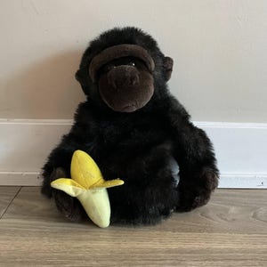 May include: A plush gorilla toy with dark brown fur and a brown face. The gorilla is holding a yellow and green banana. The toy is sitting on a wooden floor.