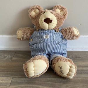 Op de afbeelding: Een pluche teddybeer met bruine vacht, crèmekleurige poten en een crèmekleurige snuit met een bruine neus. De beer draagt een blauw-witte gestreepte overall met goudkleurige knopen. De voet van de beer heeft een handtekening.