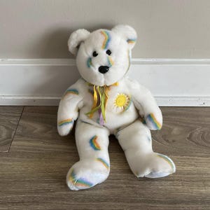Könnte beinhalten: Weißer Plüschteddybär mit Regenbogen- und Sonnenmotiv. Der Bär hat ein gelbes Band um den Hals.