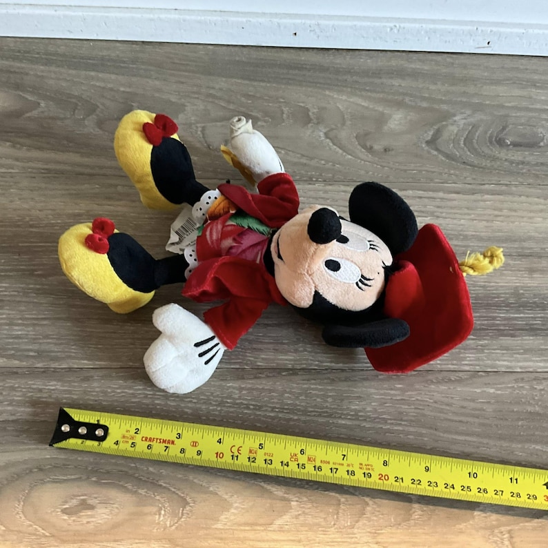 May include: A Minnie Mouse plush toy with a red dress, black ears, and yellow shoes with red bows. The toy is lying on a wooden surface next to a Craftsman measuring tape. The tape measures up to 30 inches.
