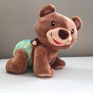 May include: A brown plush bear toy wearing a green and white nappy with a red ladybug on its back. The bear has a smiling face with a pink nose and mouth.