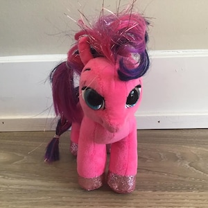 May include: Pink plush unicorn toy with purple and pink hair, sparkly hooves, and large blue eyes.