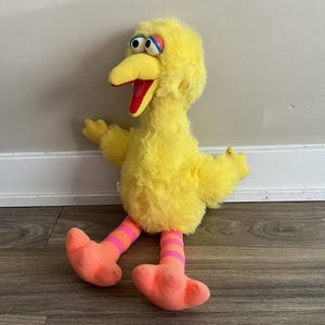 Może przedstawiać: Pluszowa zabawka przedstawiająca postać Big Bird, z jasnym żółtym futerkiem, dużym żółtym dziobem z czerwonym wnętrzem oraz szerokimi oczami z niebieskimi powiekami. Figurka ma pomarańczowo-różowe pasiaste nogi i duże pomarańczowe stopy.