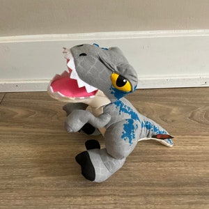May include: A gray and blue plush dinosaur toy with a large open mouth and yellow eyes. The dinosaur is sitting on a wooden floor.