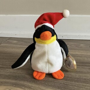 May include: A white and black penguin plush toy wearing a red Santa hat with a white pom-pom. The penguin has a yellow beak and orange feet. The toy has a Ty heart-shaped tag attached.