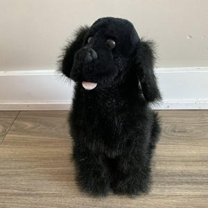 Könnte beinhalten: Ein schwarzes Plüschhundespielzeug mit Schlappohren und einer rosa Zunge. Der Hund sitzt aufrecht und hat eine weiche, pelzige Textur. Das Spielzeug ist so gestaltet, dass es einem Labrador Retriever oder einer ähnlichen Rasse ähnelt.